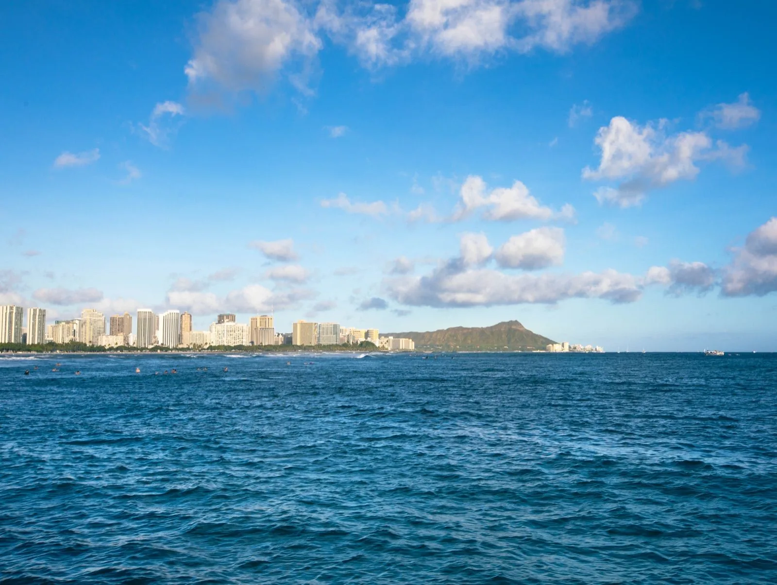 Waikīkī's high-rise shoreline and Diamond Head across open blue water, with surfers in the water and a partly cloudy sky