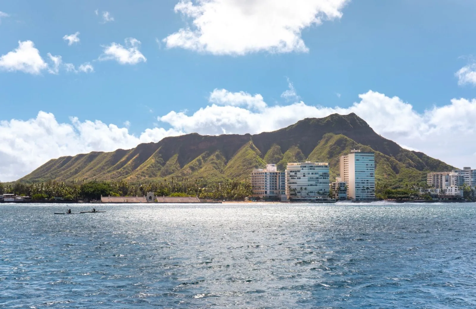 Diamond Head rising behind shoreline high-rises and palms, seen across open blue water with two paddlers in daylight