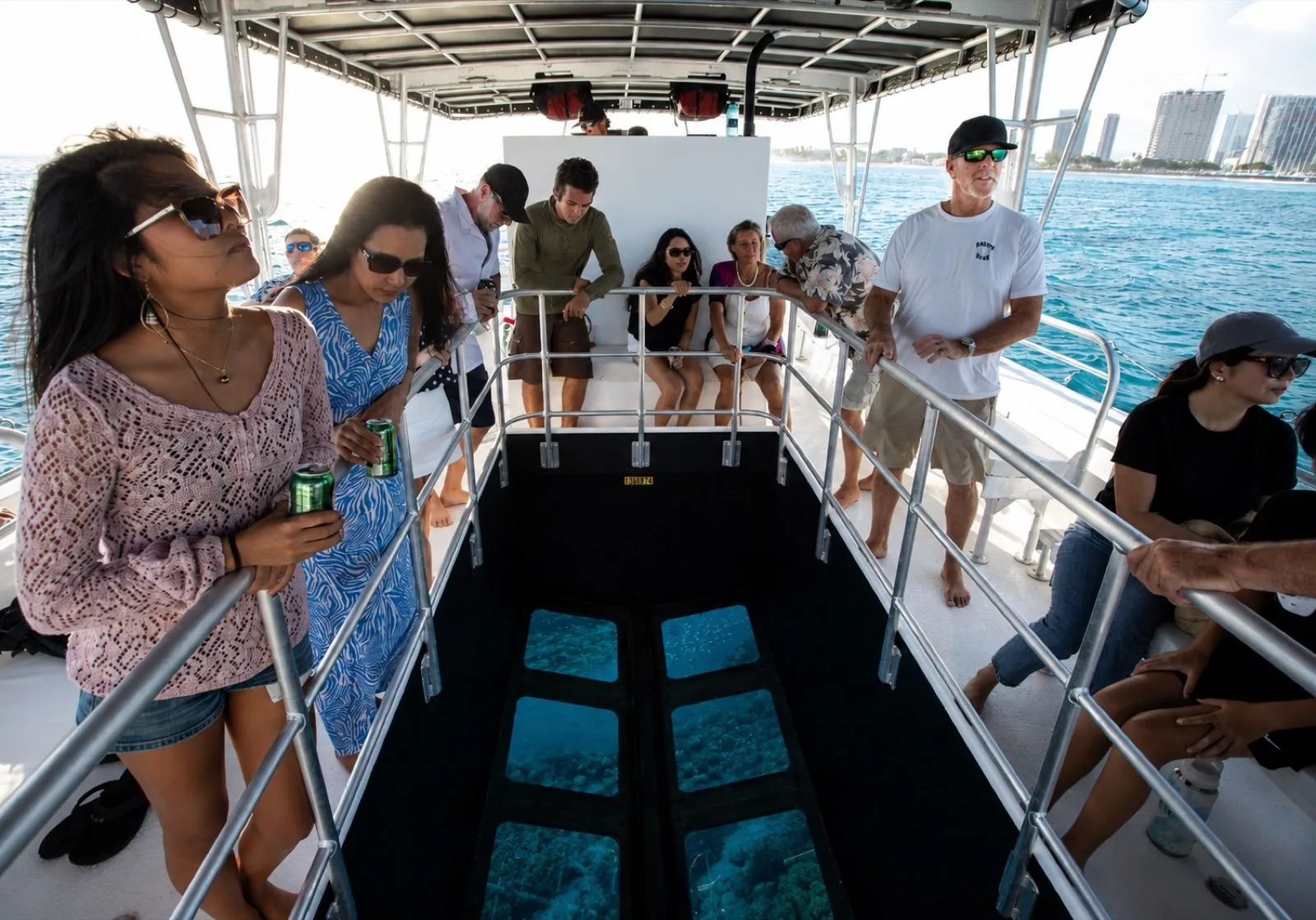 Guests stand at a metal railing around a glass-bottom viewing well on a covered boat deck, looking down at reef through the panes, with high-rises along the shore behind them