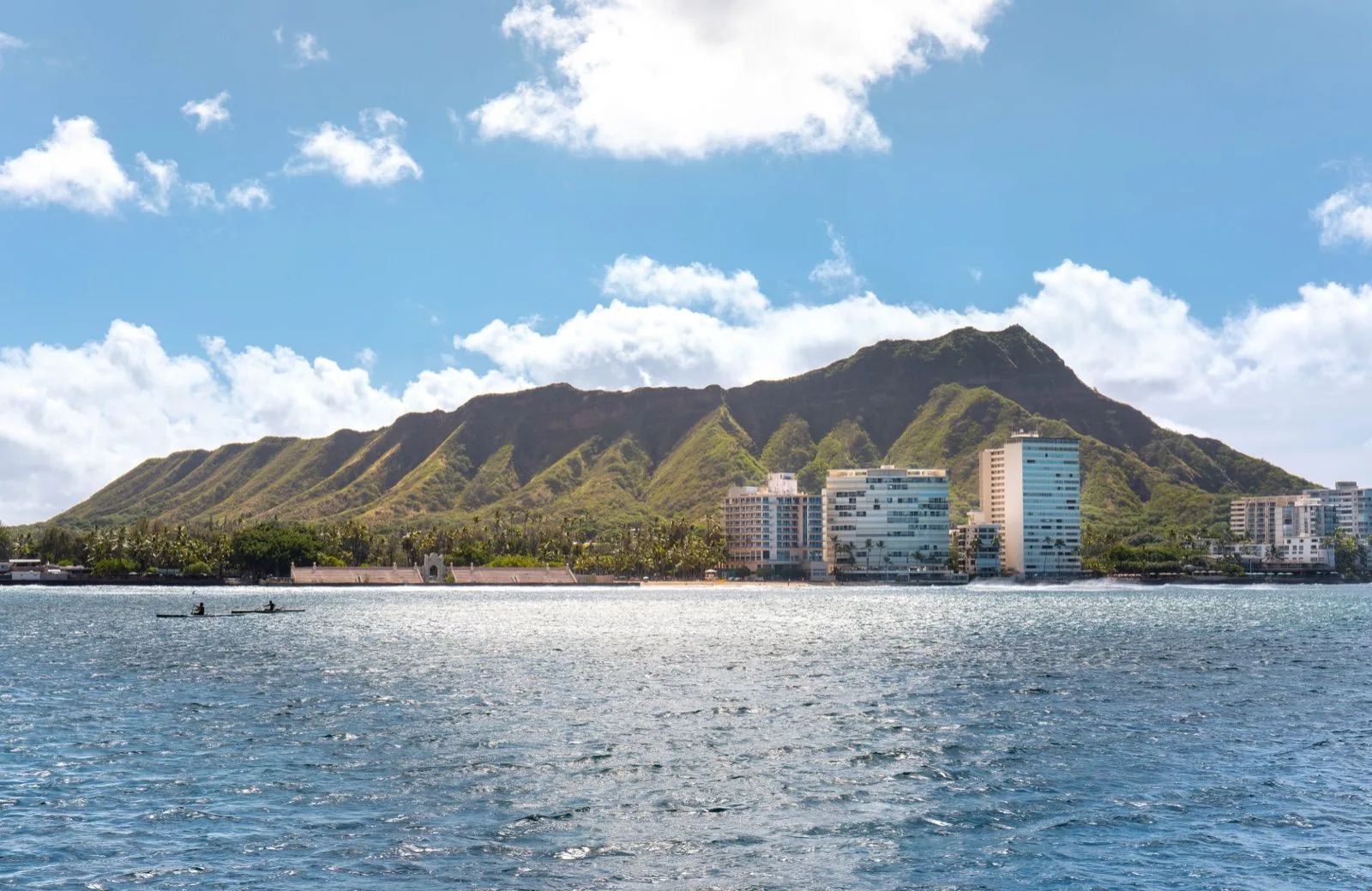 Diamond Head and Waikīkī high-rises across open water on a clear day, with two paddlers at left
