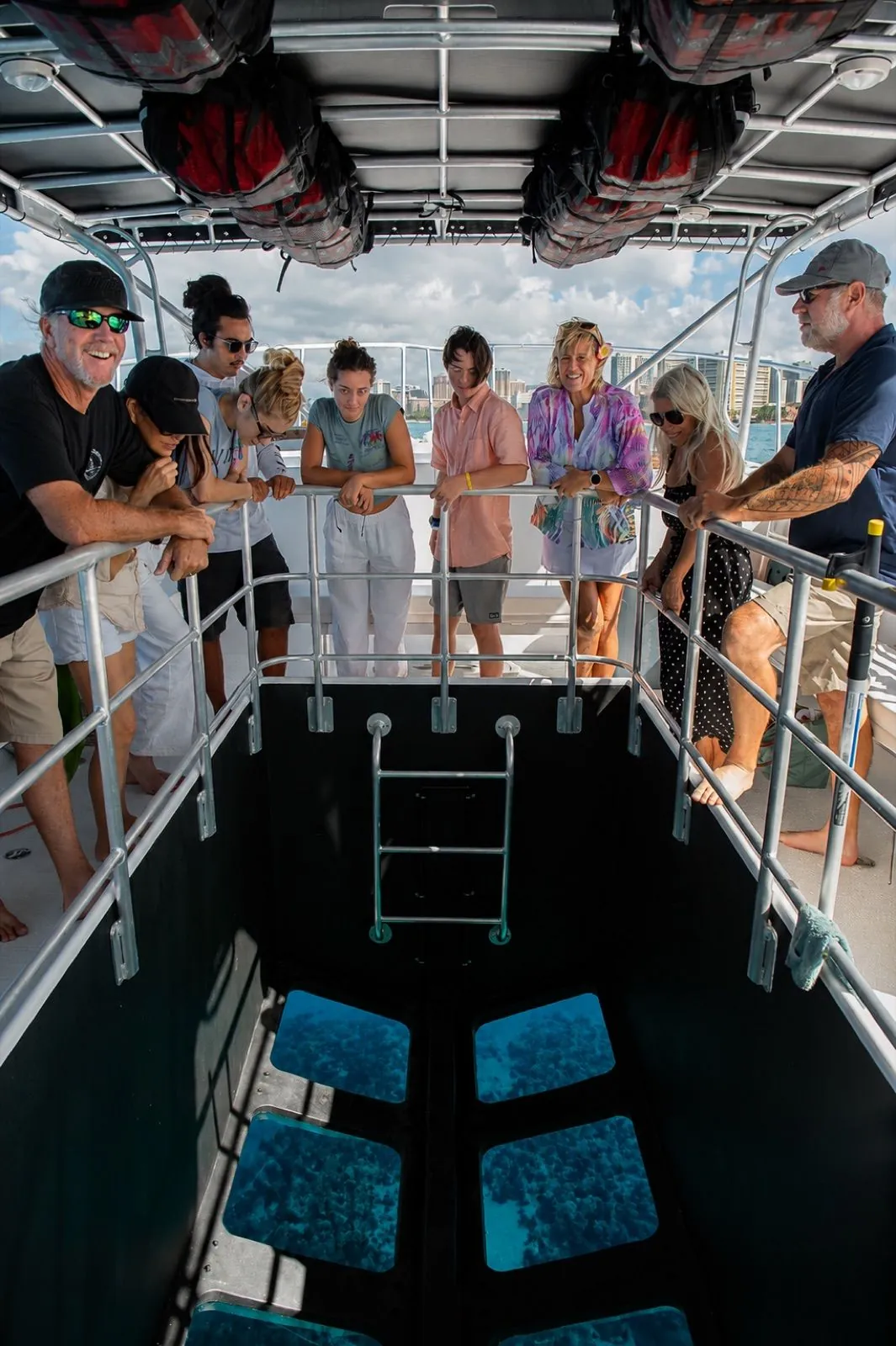 Guests lean on the rail around a glass-bottom viewing well and look down at coral through the glass panes, with high-rise buildings on the shore behind them