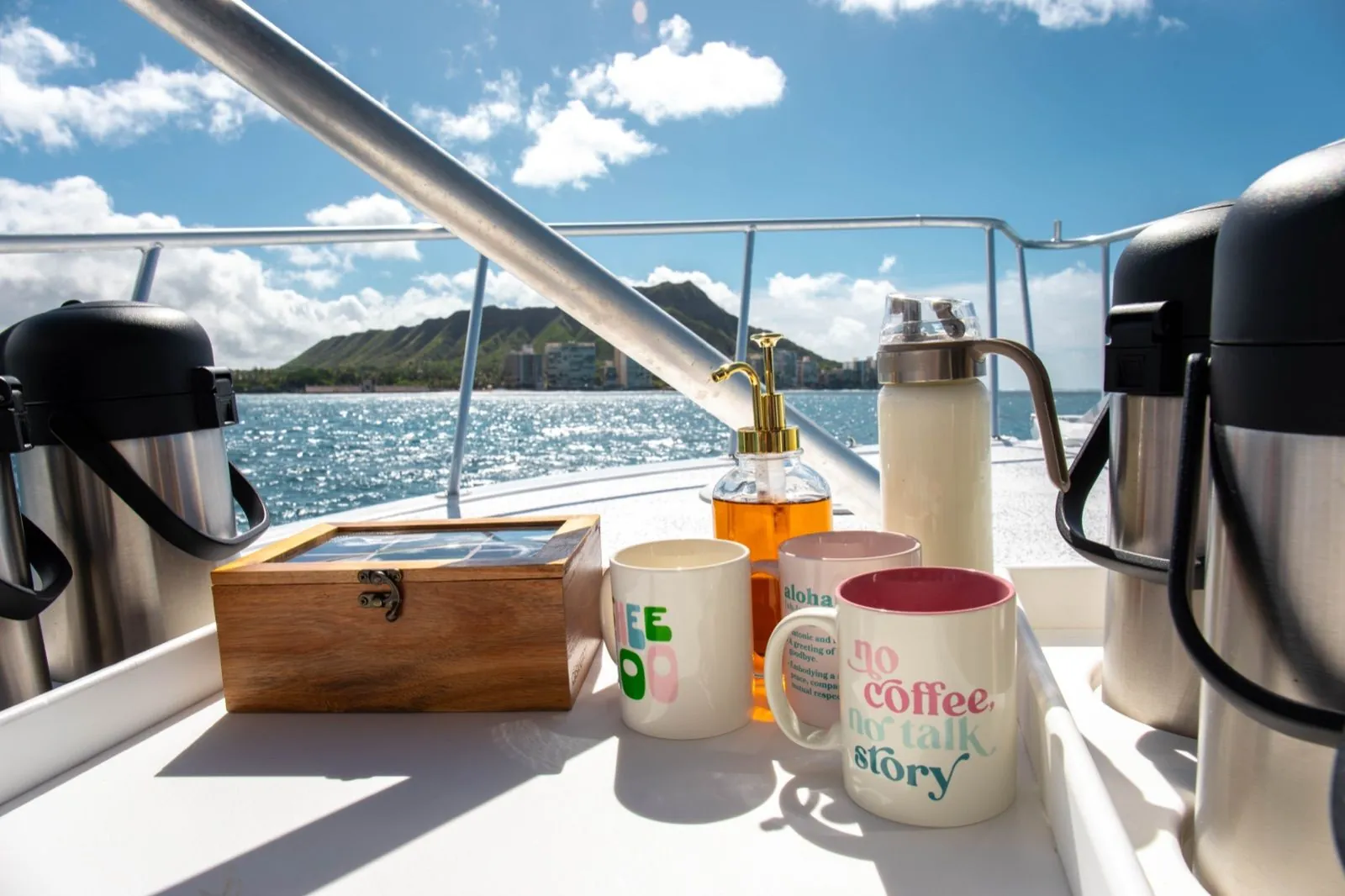 Coffee mugs, insulated flasks, a syrup pump bottle and a wooden box on a boat's serving counter, with Diamond Head and the shoreline across the water