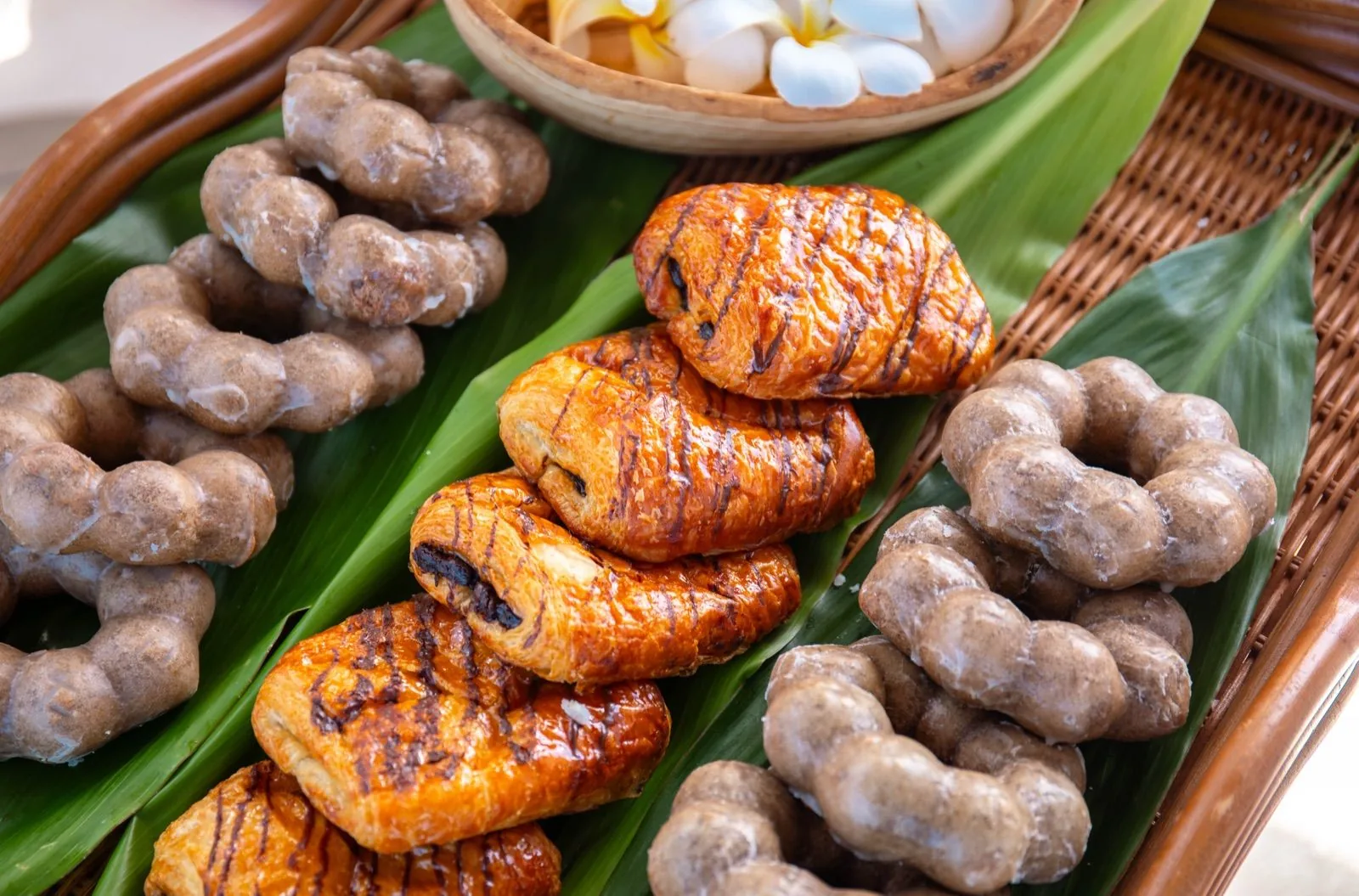 Chocolate-drizzled pastries and ring-shaped glazed pastries laid on green leaves in a rattan tray