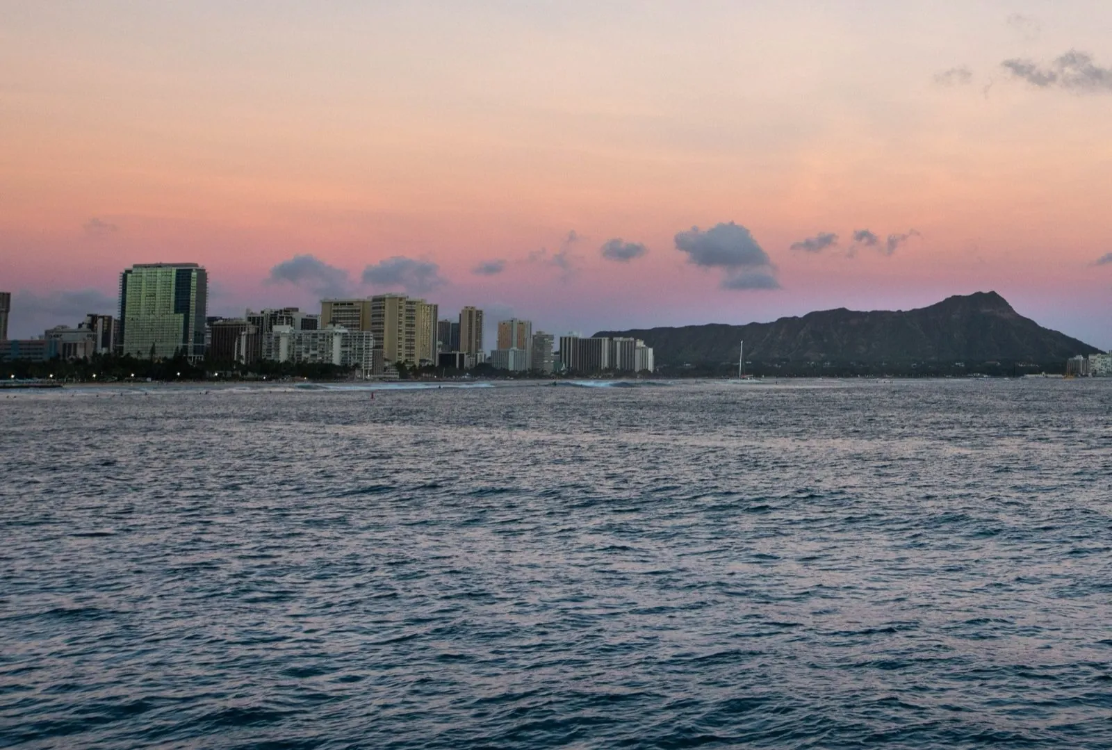 Waikīkī's hotel skyline and Diamond Head seen across open water at dusk, under a pink and lilac sky
